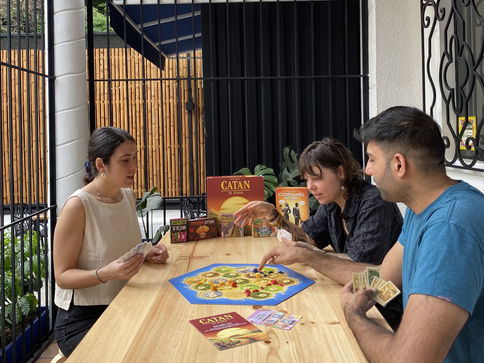Three guests around a game of Catan on the front terrace, cards in hand, with the Catan and Codenames boxes stood up beside the board and the ironwork behind them.