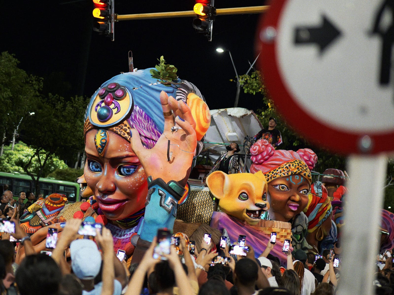 A giant painted float figure in a blue headwrap waving over the crowd at a night parade of the Feria de Cali, dozens of phones held up under the traffic lights.