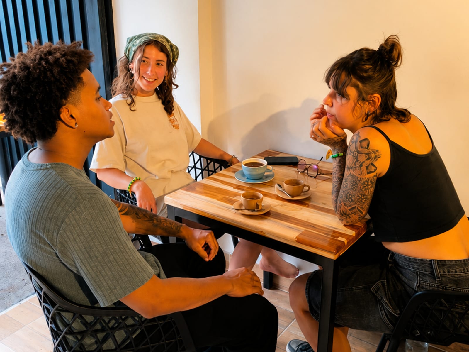 Three guests talking over coffee at a small wooden table in the café, three cups, a phone and a pair of glasses between them, one resting her chin on a tattooed arm.