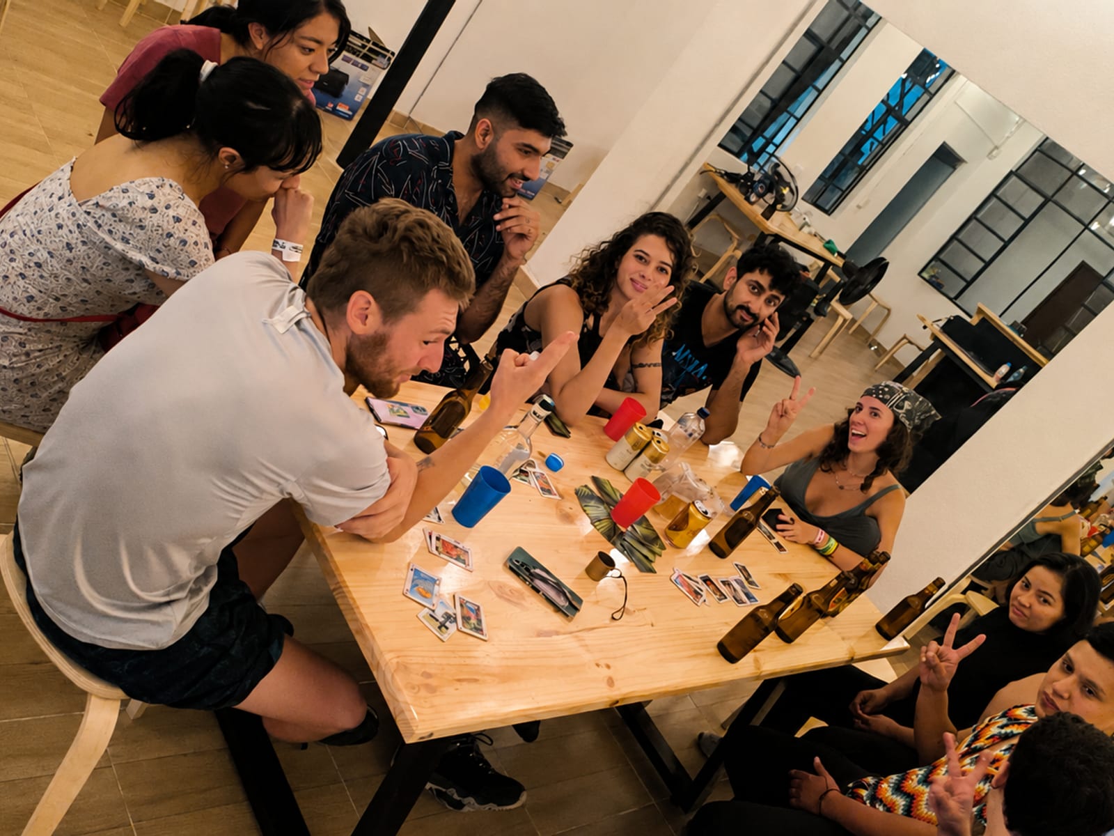 About ten guests around the long wooden table at night, cards and beer bottles between them, one mid-gesture and several laughing, the dark window and a mirror wall behind.