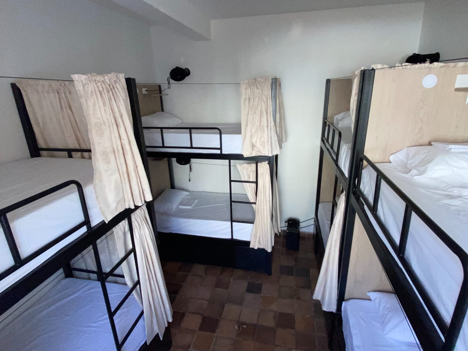 A six-bed mixed dorm: the far corner, a made bunk with its curtain drawn back and a reading light on the wall.